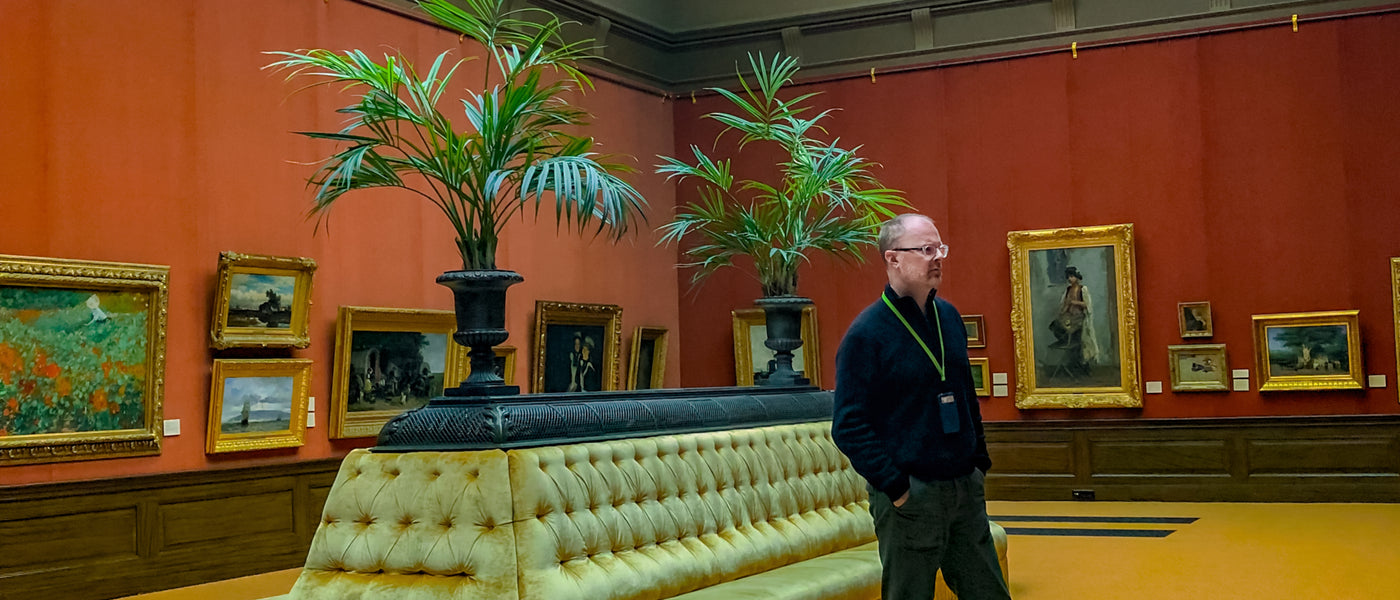 Photo of Christopher St.Leger in an art gallery looking at impressionist paintings. Gallery has terra-cotta red walls, a large tufted yellow sofa with palm plants behind it. All of the paintings have ornate gold frames.